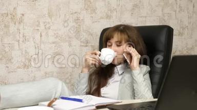 办公室里的女人在工作场所喝咖啡休息。 有电话的漂亮女商人光着脚坐在椅子上